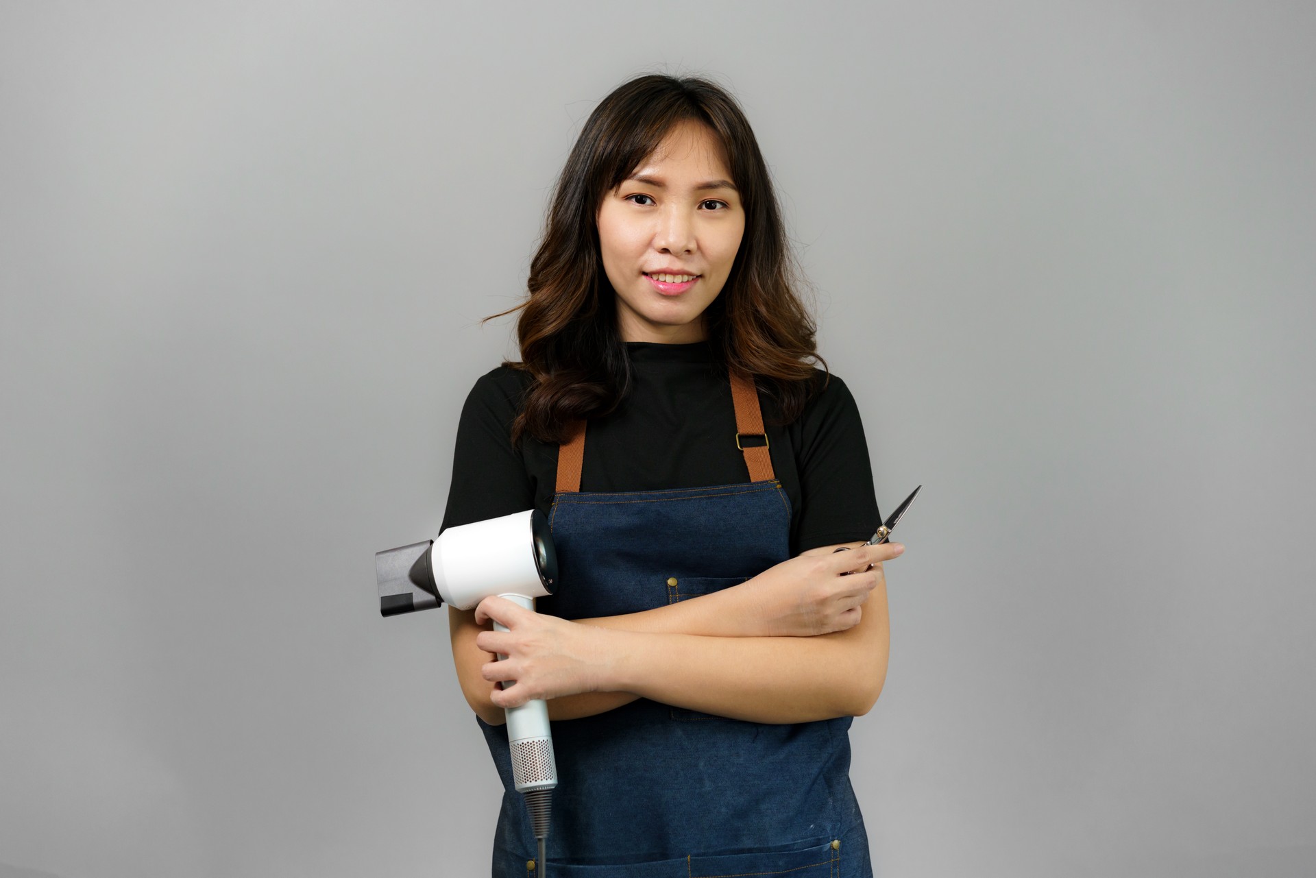 Portrait of Asian gorgeous cheerful young female professional hairdresser holding blowdryer smiling to the camera posing at her beauty studio.