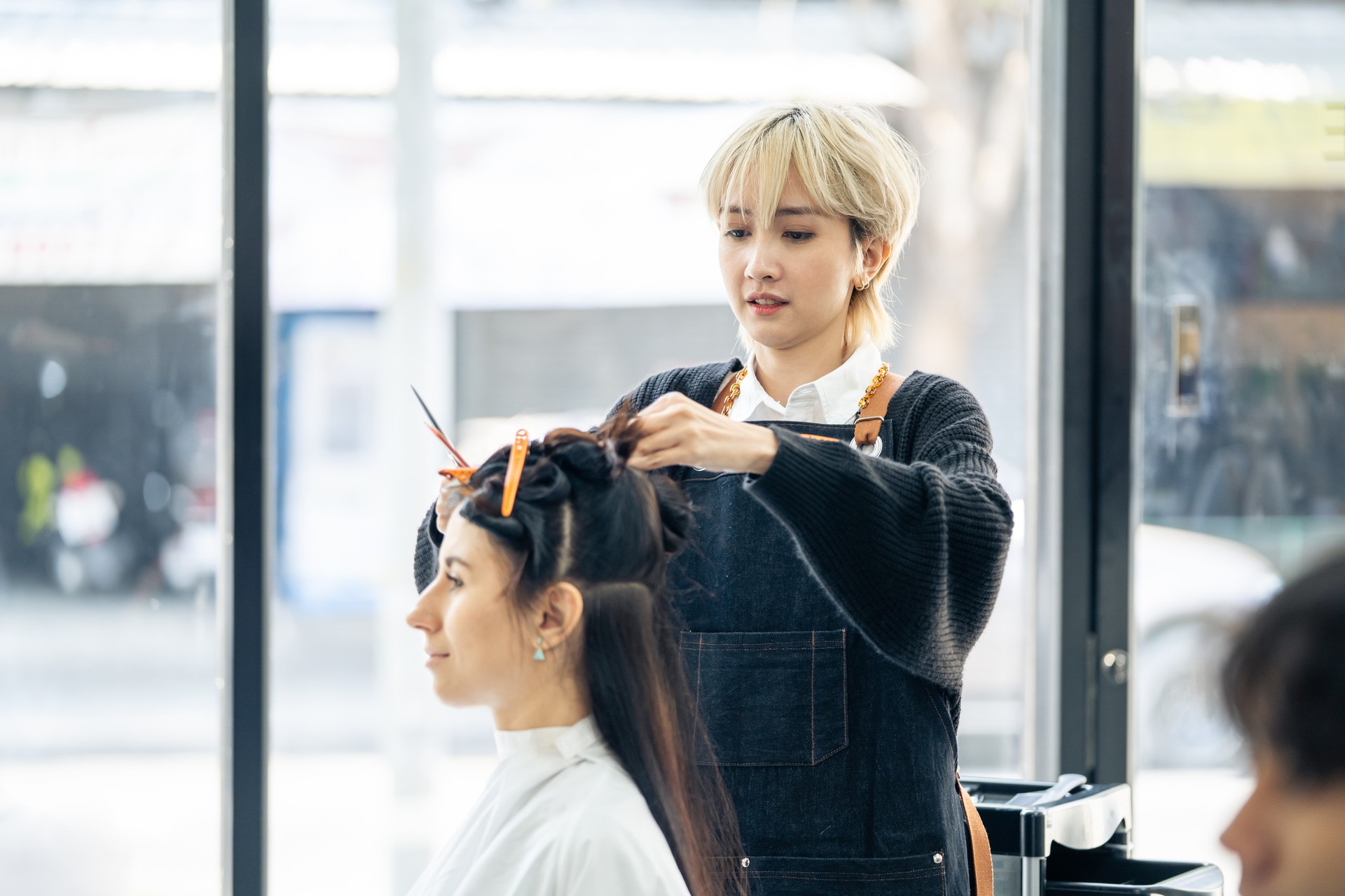 Asian professional female stylist cutting woman's hair in salon. The woman hairdresser using scissors cut the young girl sitting in the shop looking at mirror with smile. Beauty salon business concept